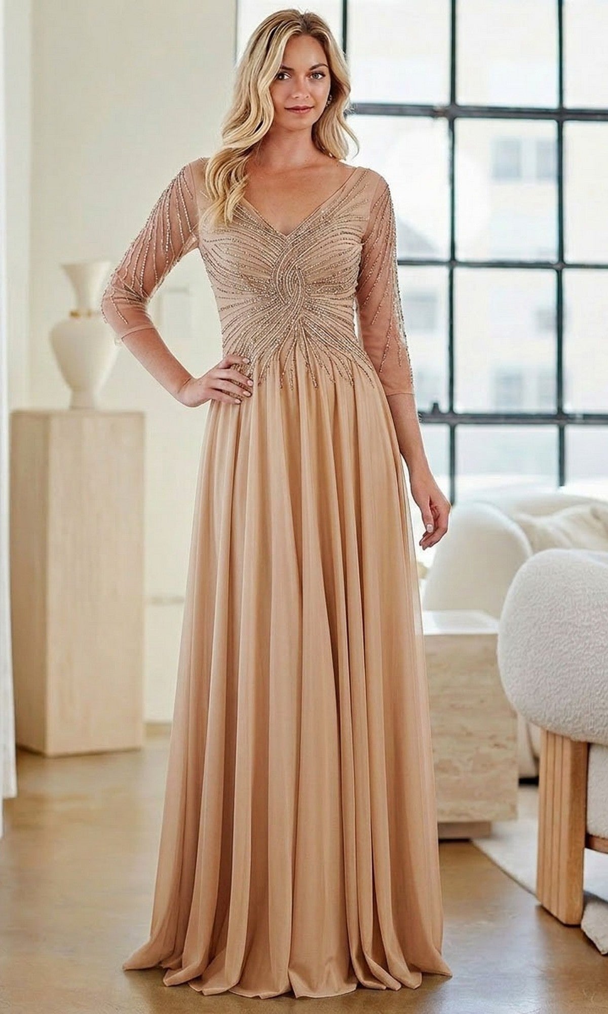 A woman is indoors wearing the gold Long Formal Dress: Amelia Couture KF1501, which features a beaded bodice, sheer embellished sleeves, and an elegant A-line skirt.
